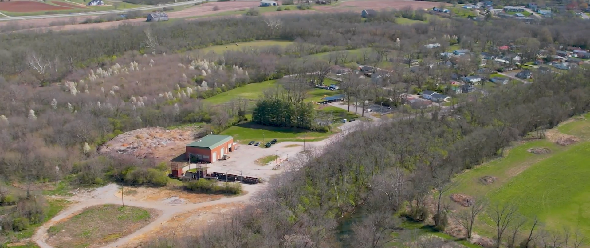 Aerial view of a building surrounded by trees and a grassy area. A small town is visible in the background.