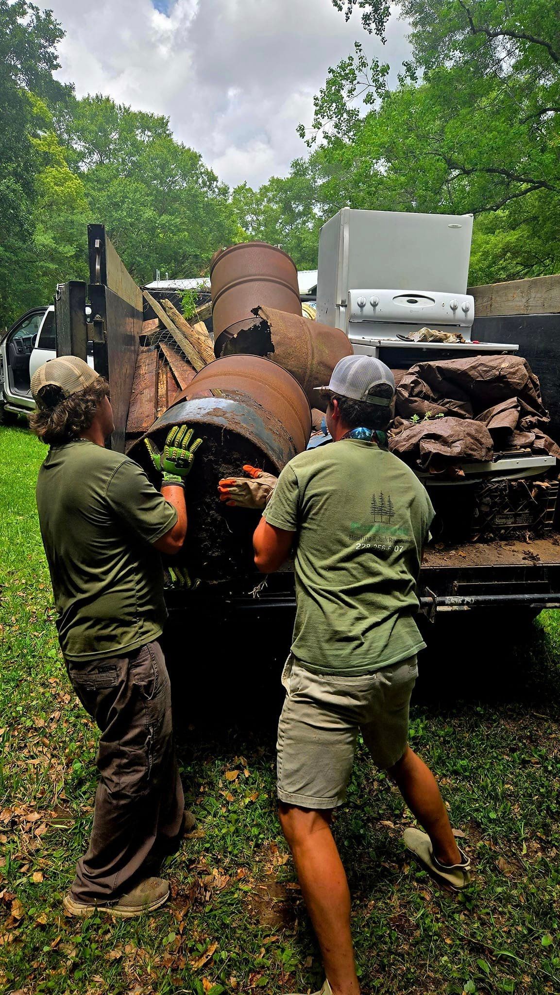 Two people loading rusty metal barrels and debris into a truck outdoors.