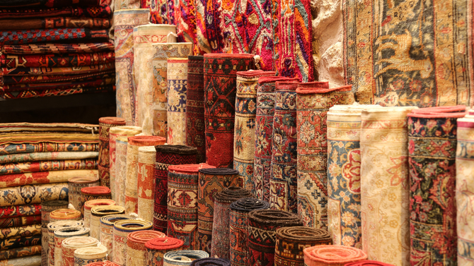 Rolled-up, colorful carpets on display in a shop; various patterns and hues, mostly reds, browns, and blues.