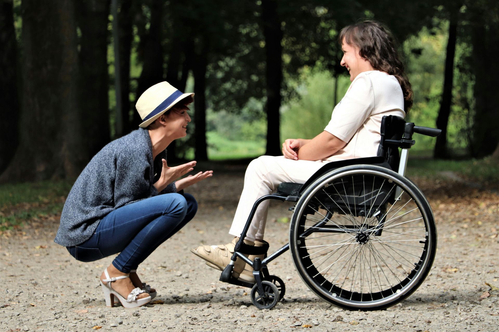 Woman crouches talking to another woman in a wheelchair on a path in a park.
