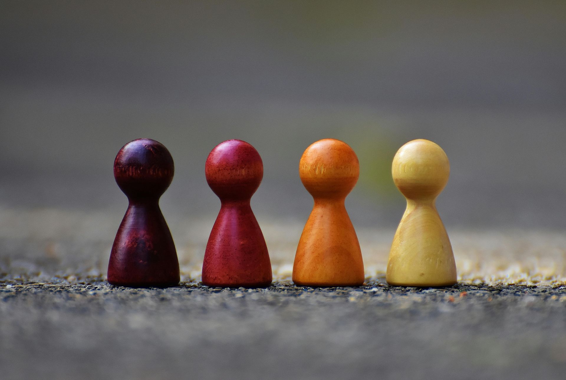 Four wooden game pieces, dark purple, red, orange, and yellow, standing in a row on a textured surface.