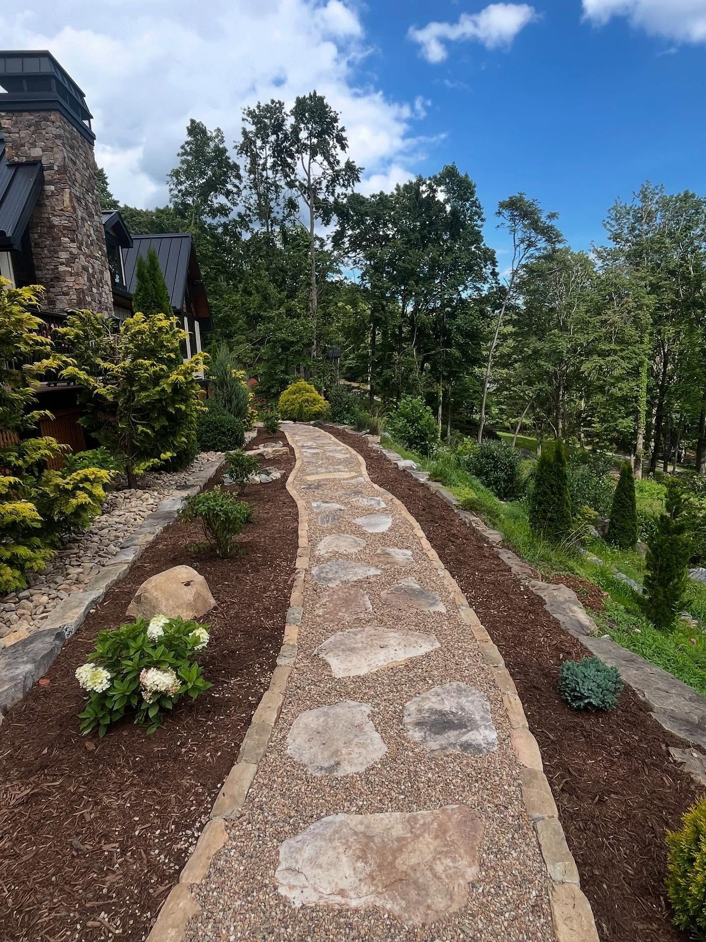 Stone path winds through a garden with trees and shrubs under a blue sky.