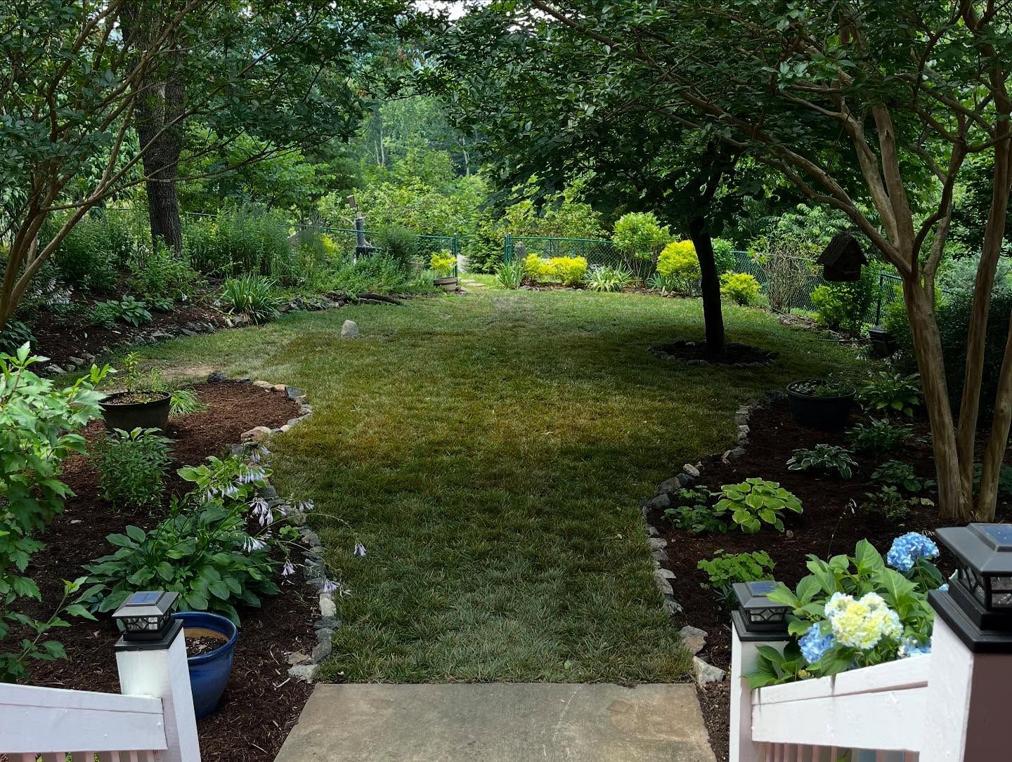Lush green backyard, viewed from a porch. Trees frame a grassy area with garden beds, flowers, and a path.