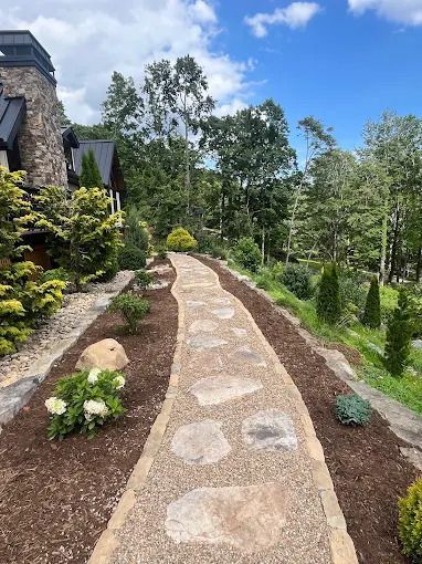 Stone pathway winding through a landscaped yard with trees and a house on a sunny day.