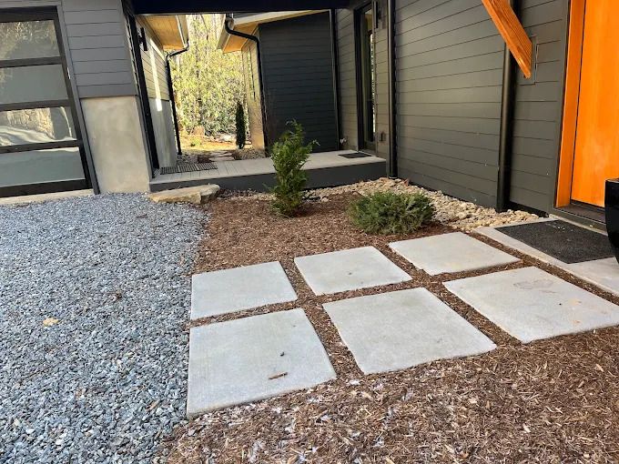 Exterior entrance with gray siding, a gravel area, concrete stepping stones, and a small shrub.
