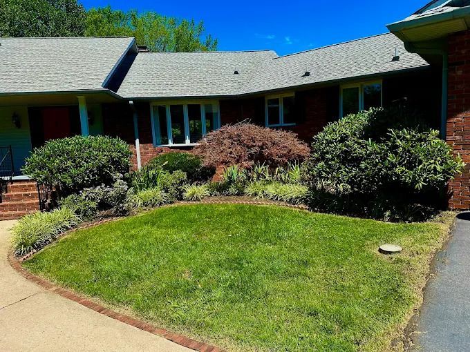 A well-manicured lawn with brick-bordered garden beds in front of a brick house with a gray roof and windows.