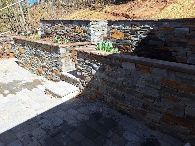 Stone retaining walls with brown coping and a patio in a yard.