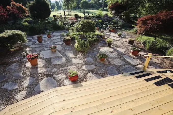 Stone patio with potted plants, surrounded by greenery, viewed from a wooden deck.