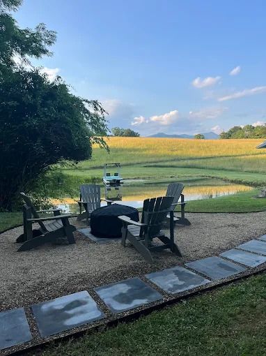 Lounge chairs around a fire pit overlooking a pond and field with mountains under a cloudy sky.