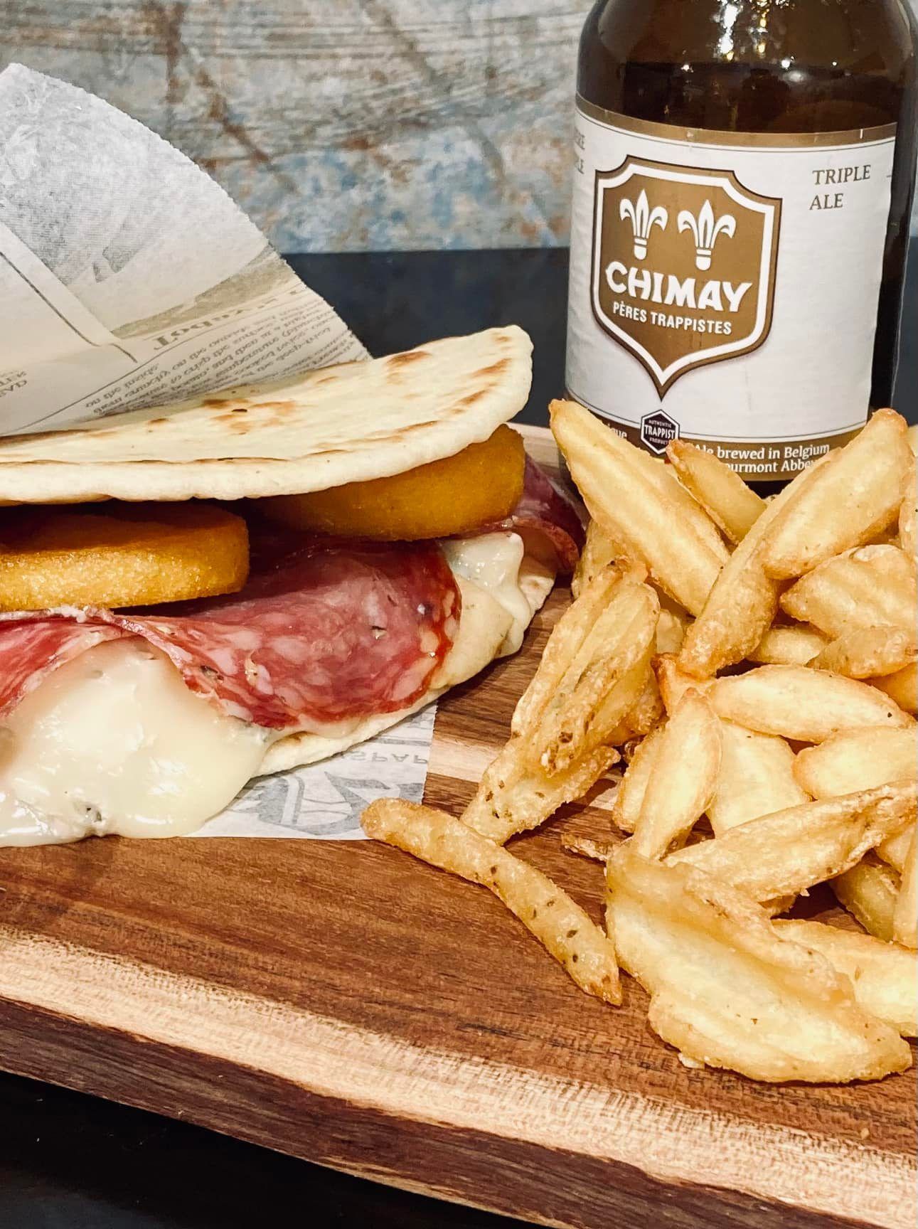 Un panino e delle patatine fritte sono su un tagliere di legno accanto a una bottiglia di birra Chimay.