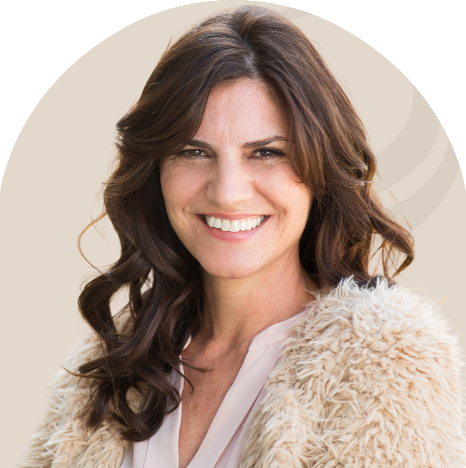 Woman with wavy brown hair smiles broadly, wearing a fluffy cream-colored coat.