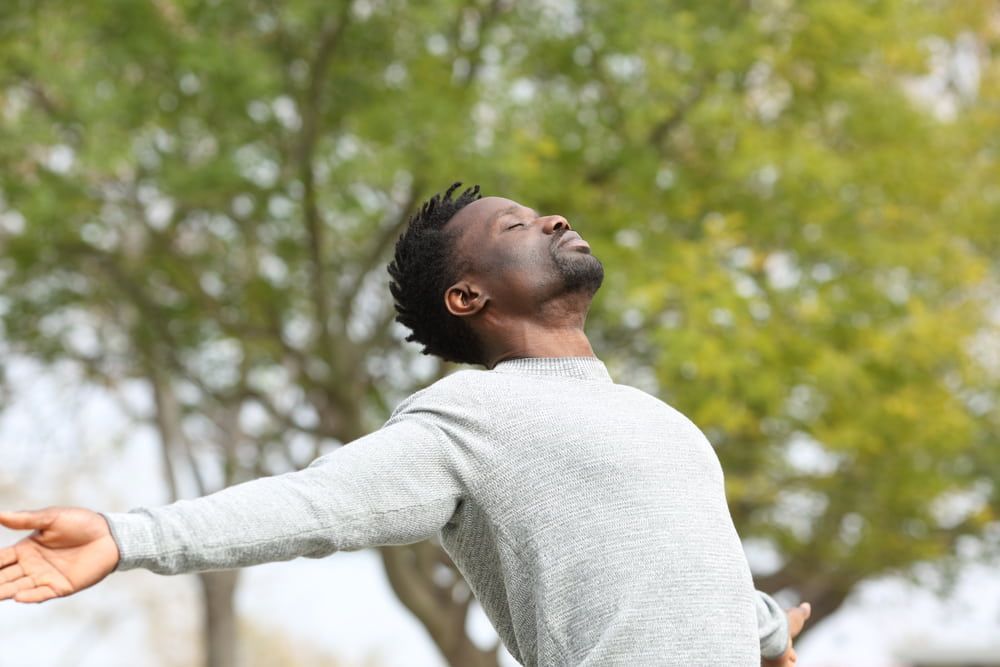 Person outdoors with arms outstretched and head tilted back near a leafy tree
