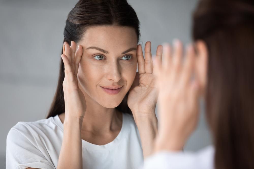 Woman looking in mirror, touching forehead, light-skinned, wearing white shirt.