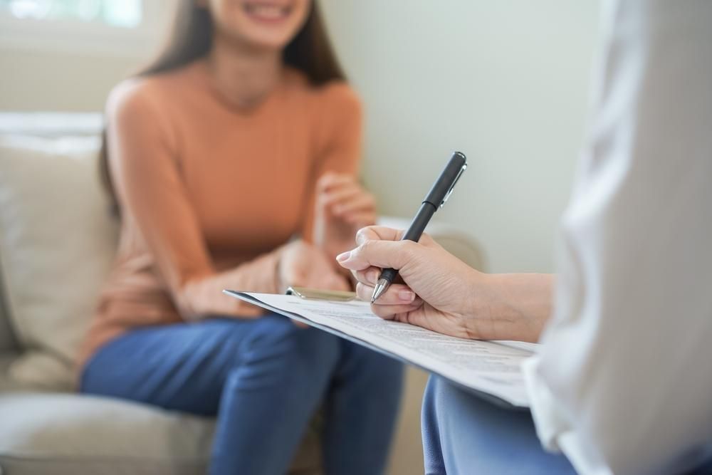 Person writing on clipboard, smiling person in background. Indoors, natural light.
