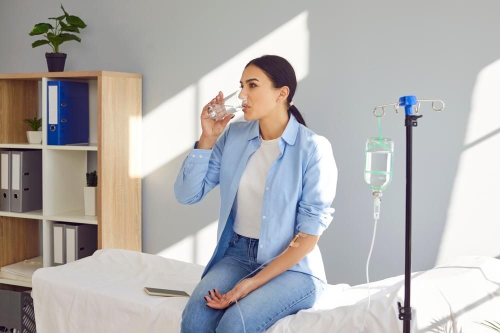 Woman drinking water while sitting on a medical exam table with an IV drip in a doctor's office.