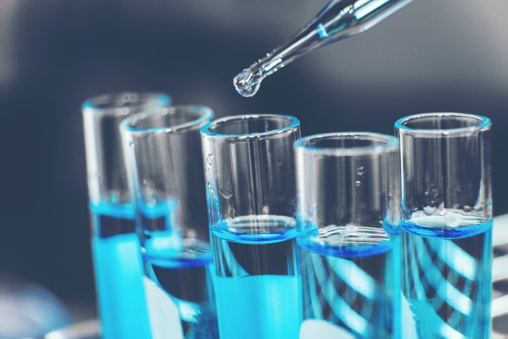 Dropper dispensing liquid into test tubes filled with blue liquid in a lab setting.