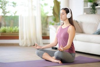 Woman meditating indoors on a purple mat, wearing headphones. 