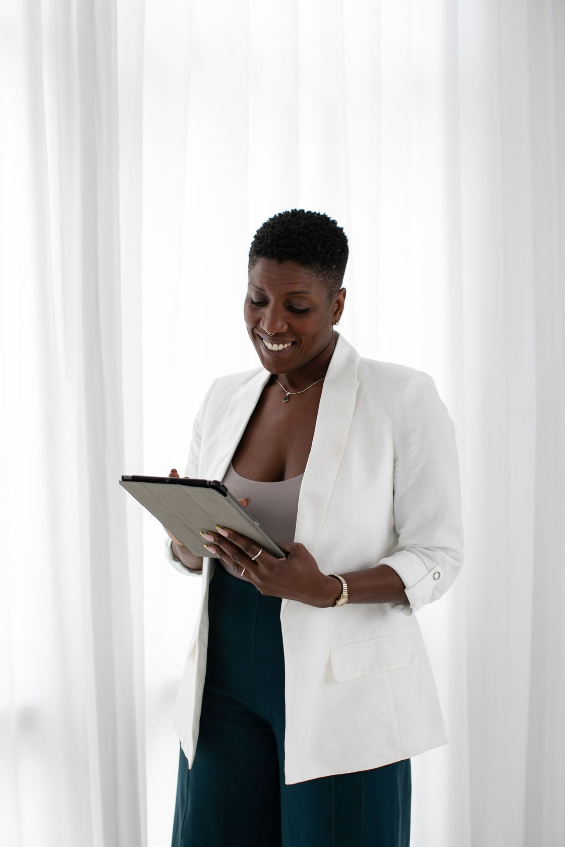 Person in a white blazer smiling while looking at a tablet in front of sheer curtains