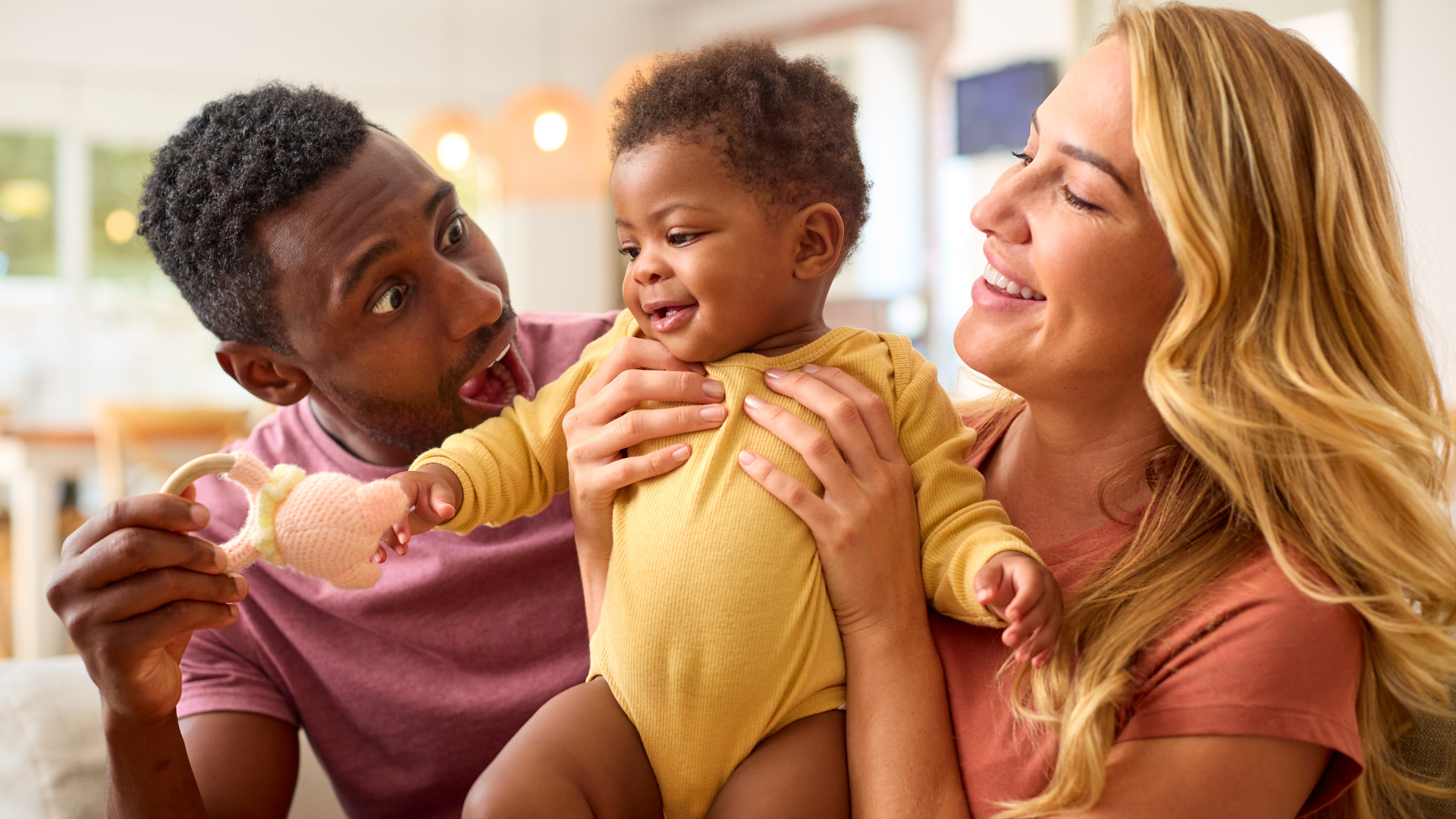 Parents smiling, holding and playing with baby, who is also smiling, indoors.