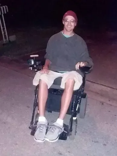Man in wheelchair wearing a red hat, olive shirt, khaki shorts, and grey sneakers outside at night.