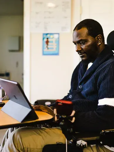 Man in wheelchair using a tablet, indoors. He's wearing a blue sweater, smiling, and has brown skin.