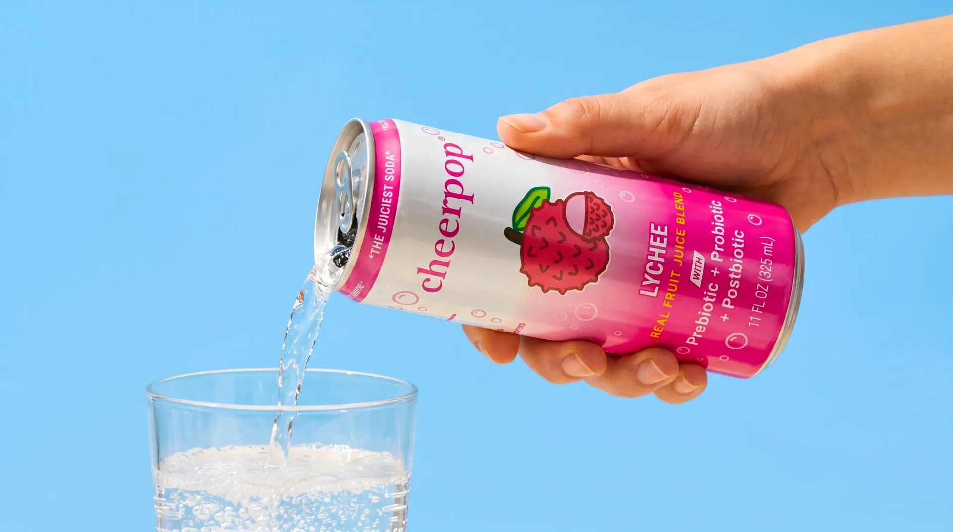 Hand pouring a pink beverage can into a clear glass against a blue background