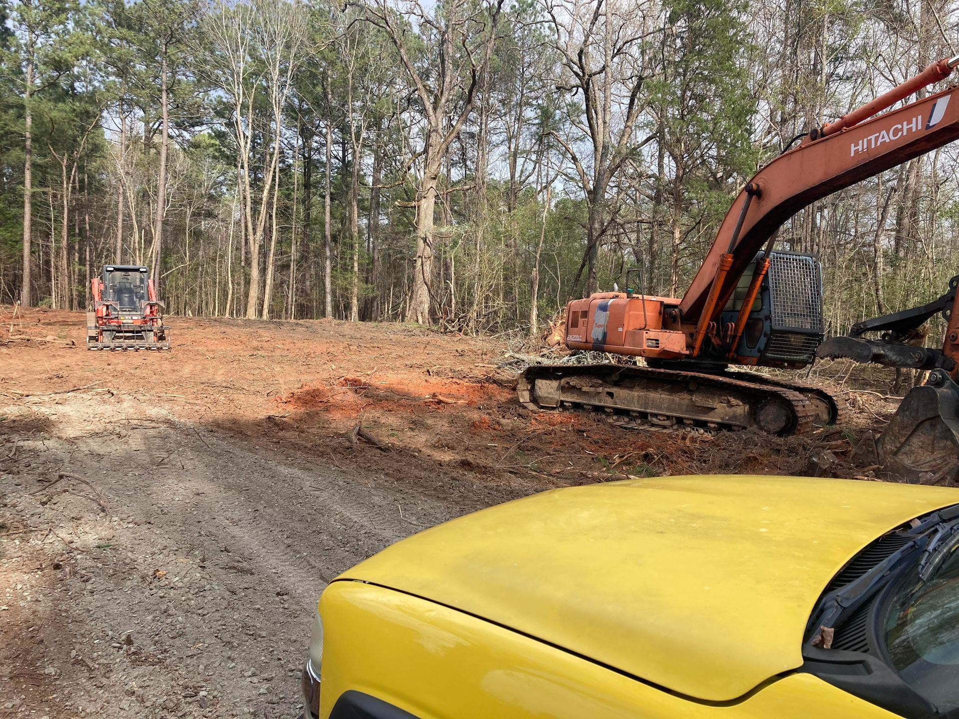 A view from a yellow truck showing an orange excavator and a small loader at a wooded construction site with cleared dirt.