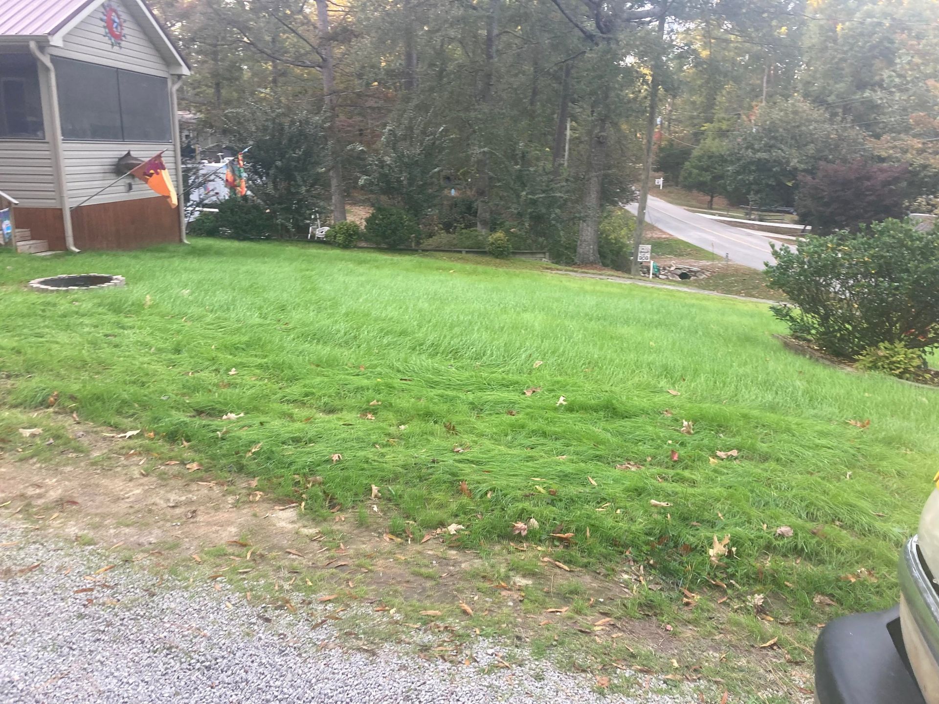 A sloped, grassy lawn in front of a small house with a screened porch, bordered by gravel and trees.