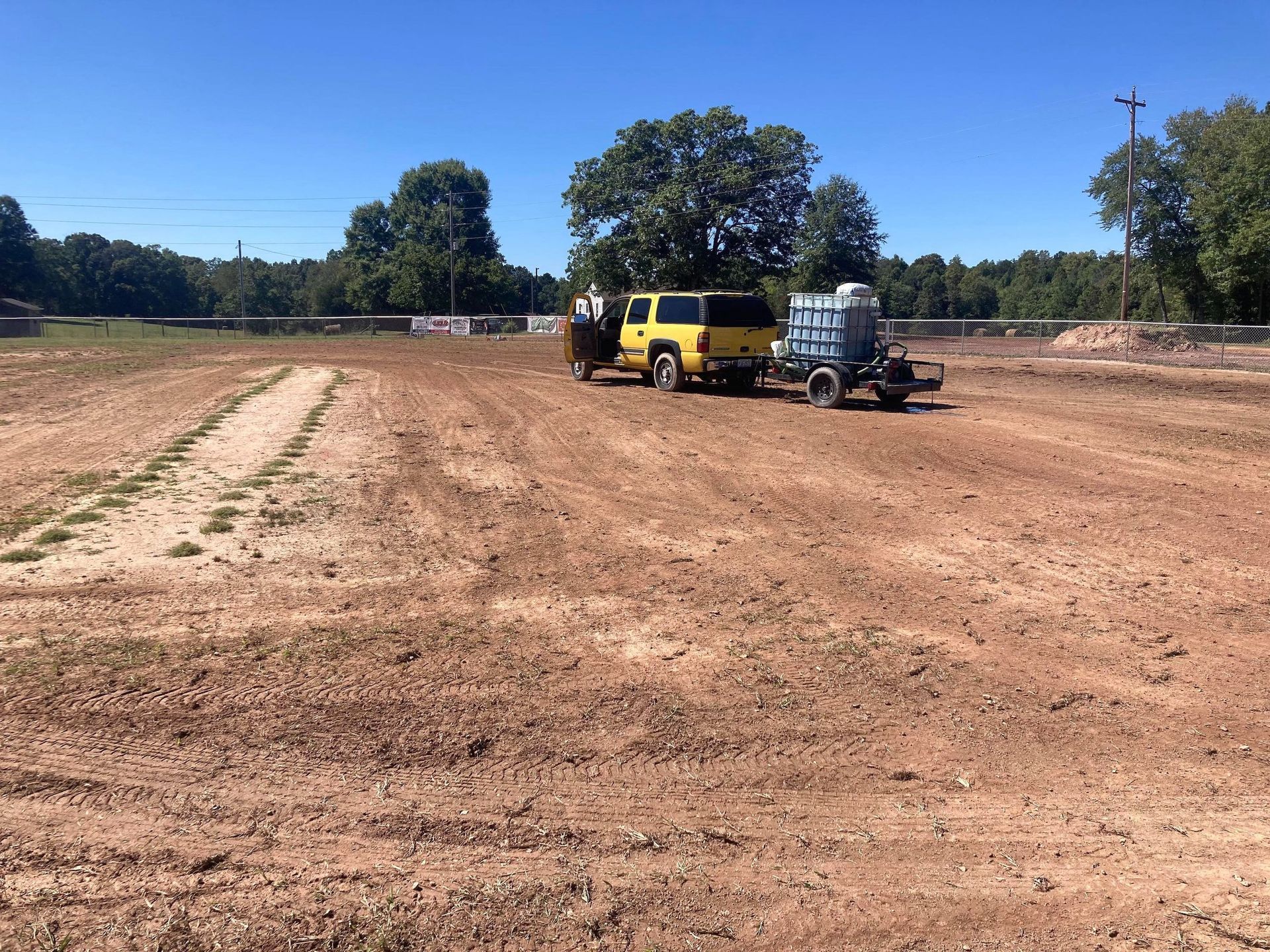 A yellow pickup truck with a trailer parked in the middle of a large, cleared dirt construction site under a blue sky.