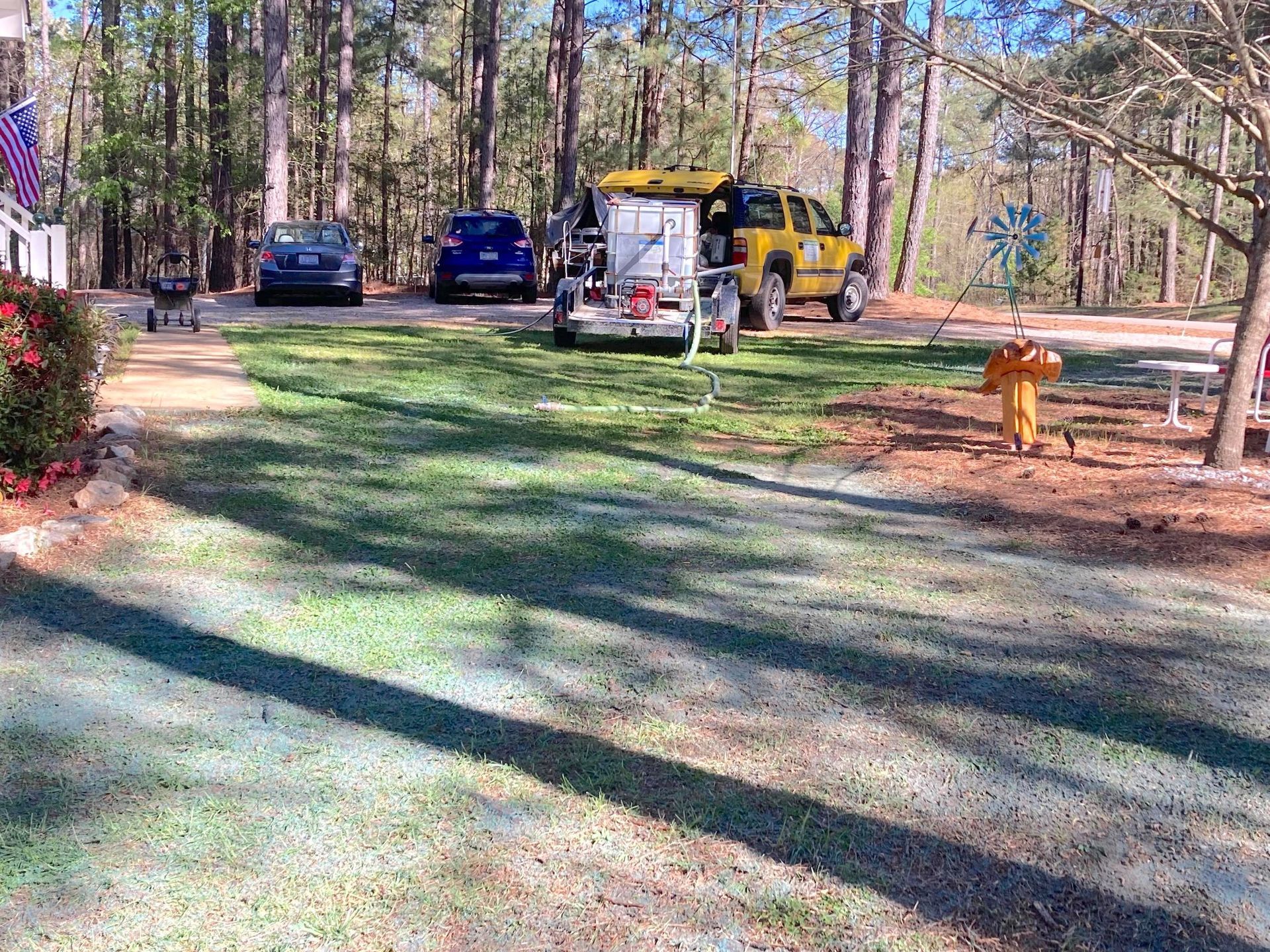 A yellow utility truck with a trailer parked on a grassy yard next to cars, with pine trees in the background.