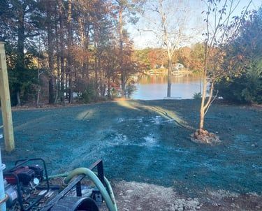 A pump sits near a patch of ground covered in blue-tinted hydroseed, located in a backyard overlooking a lake.