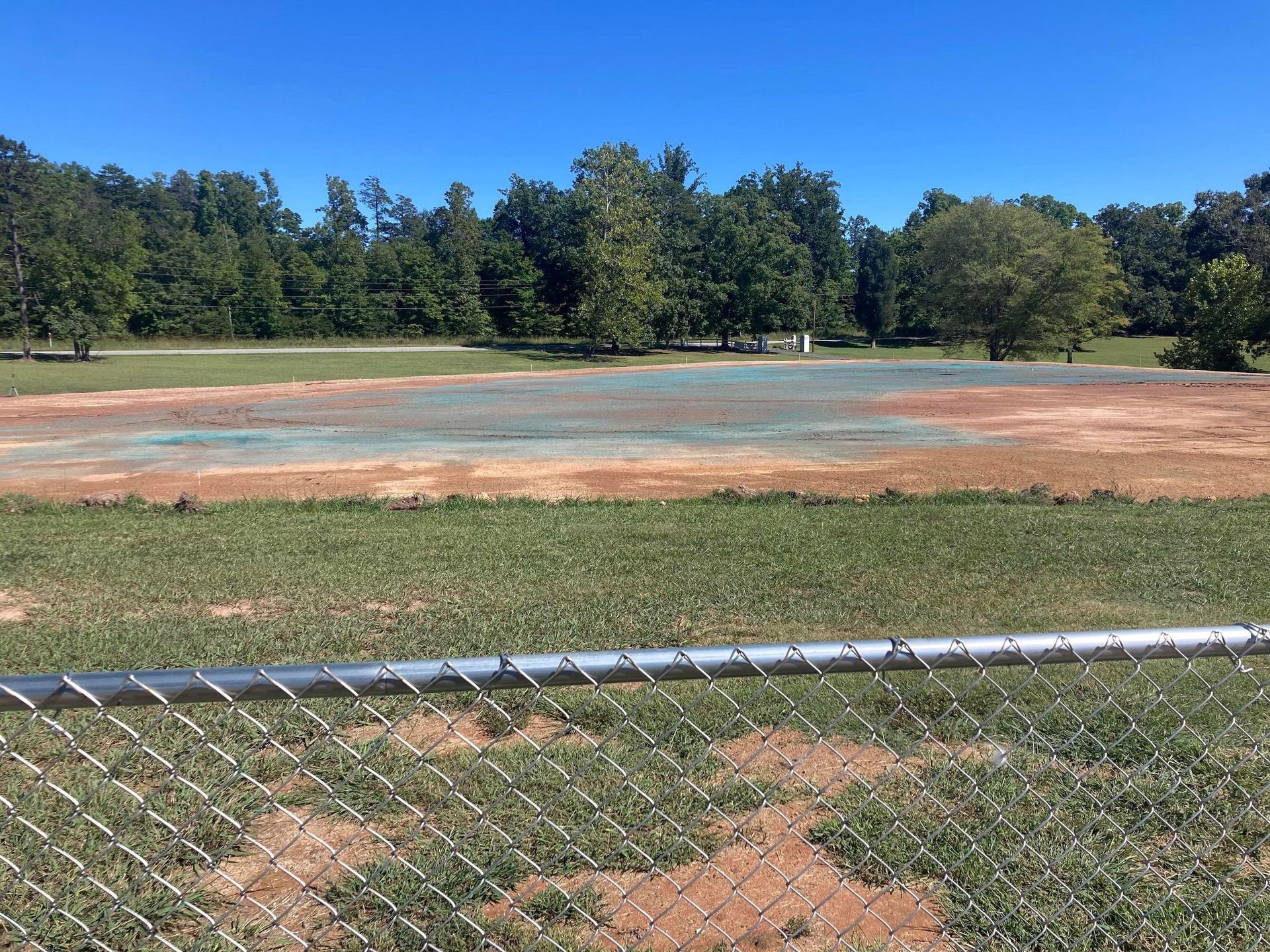 A chain-link fence in the foreground overlooks a grassy field with a large, dirt-filled area in the center under blue sky.
