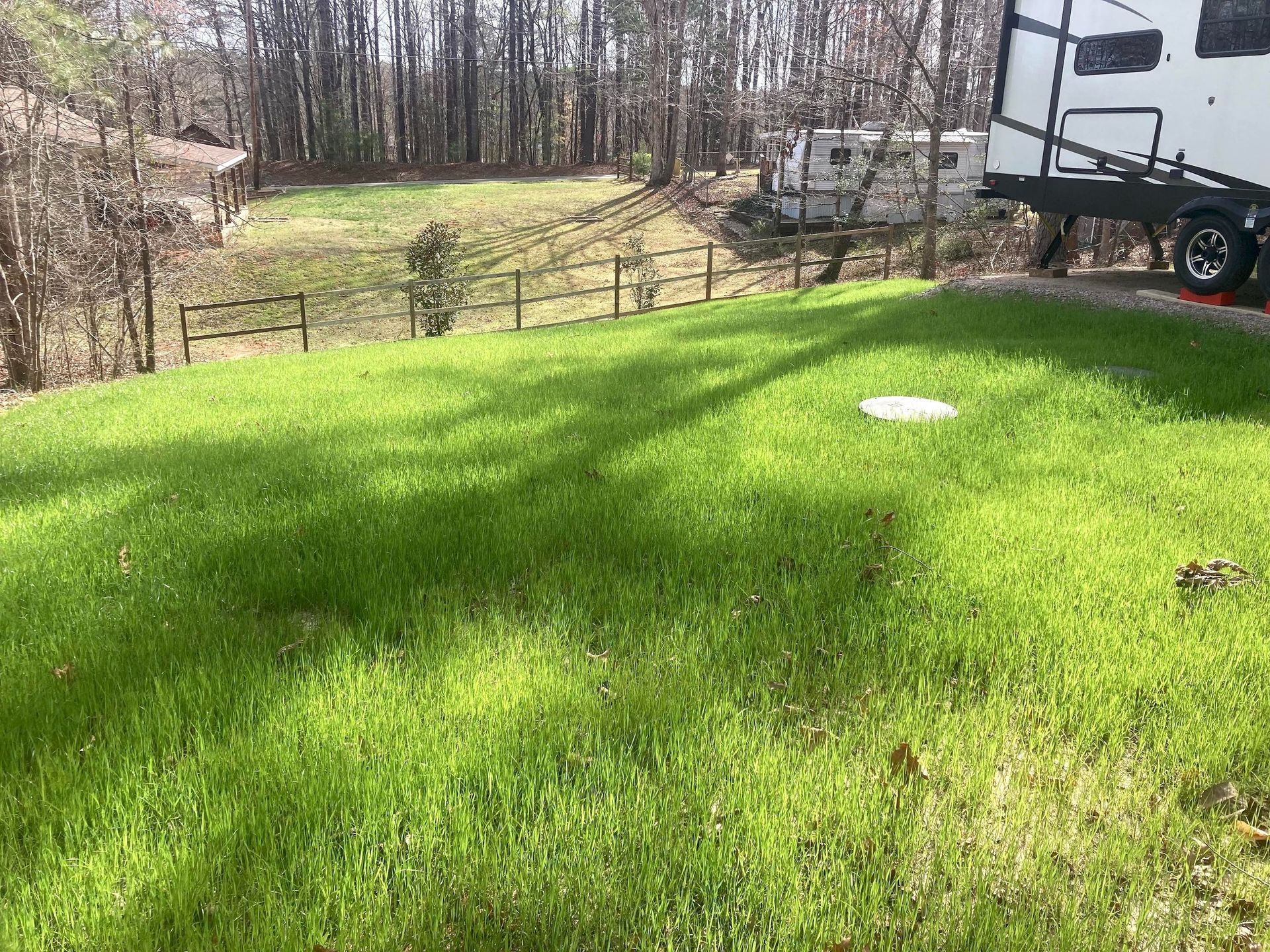 A bright green lawn leads to a wooden fence, with trees and a white camper in the background on a sunny day.
