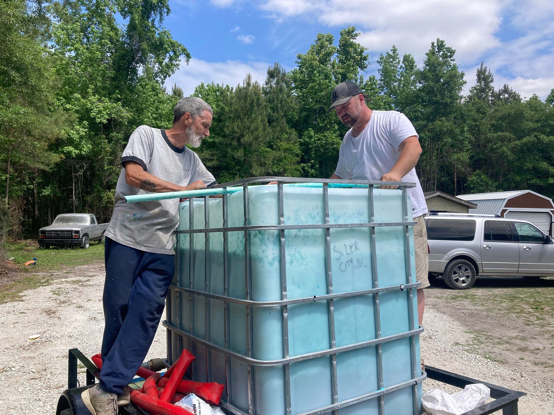 Two people stand on a trailer next to a large, light-blue water tank outdoors on a sunny day.