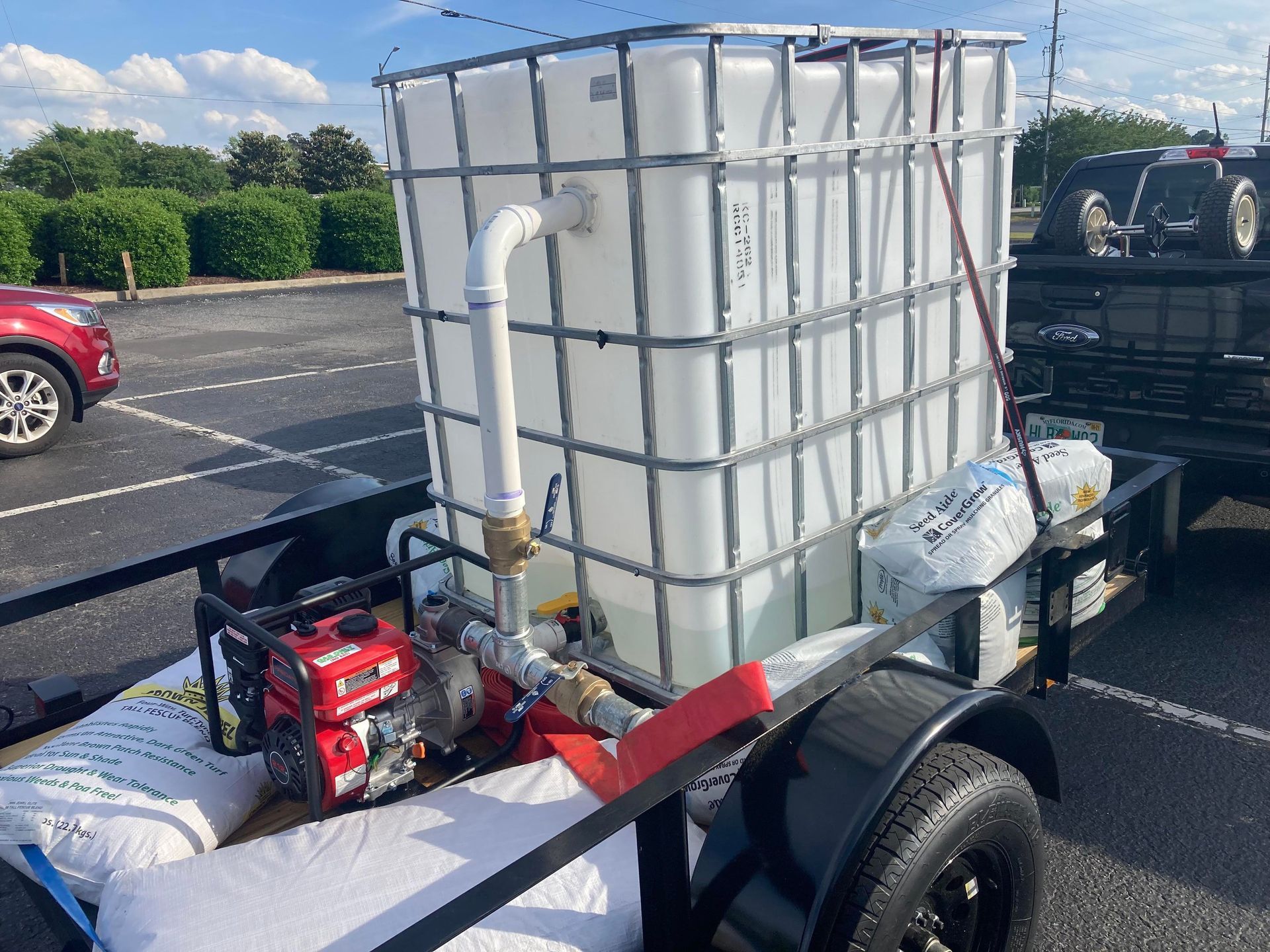 A white IBC water tank attached to a gas-powered pump on a utility trailer parked in a lot.