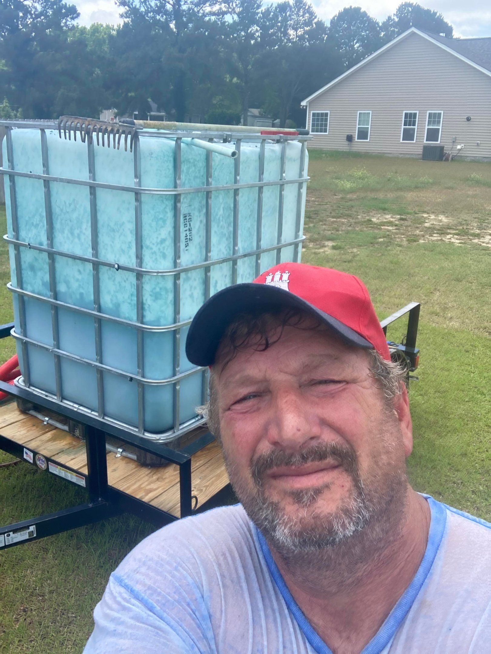 A man in a red cap poses for a selfie in front of a large, light-blue water tank mounted on a trailer in a grassy yard.