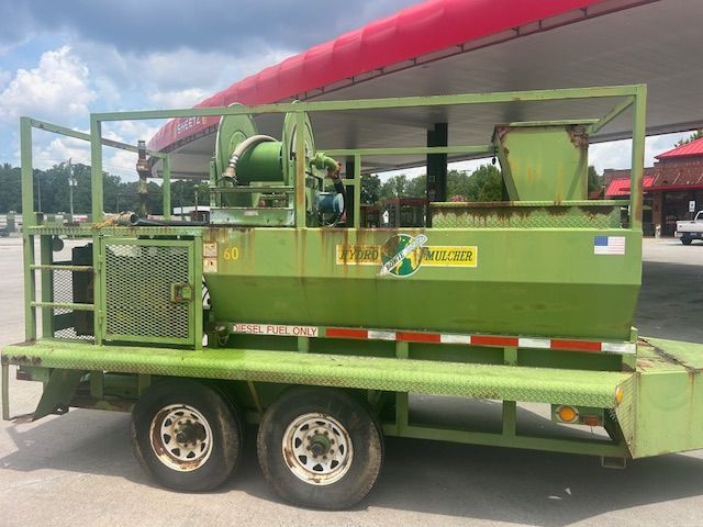 A light green industrial trailer with a mixing tank and reel, parked under a red canopy at a gas station.