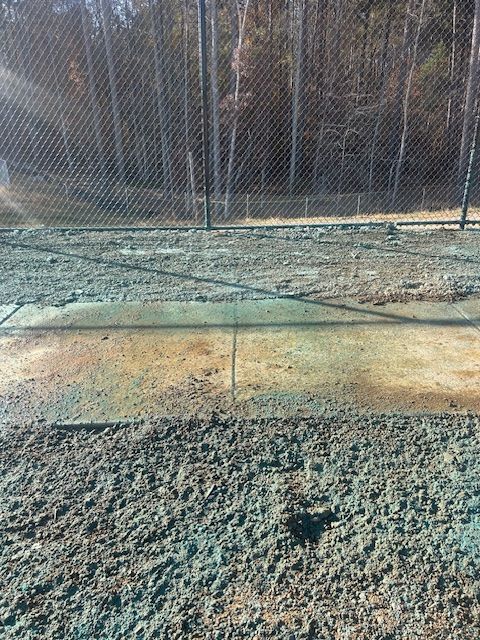 A concrete walkway sectioned by joints sits in front of a chain-link fence bordering a wooded area.