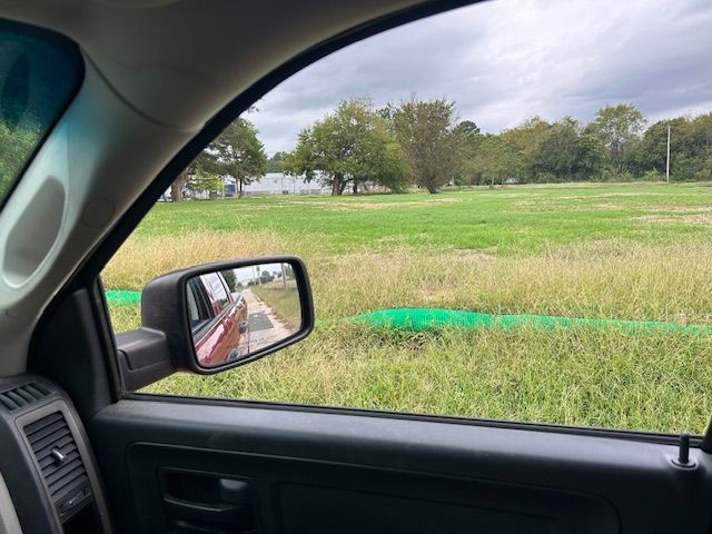 View from inside a vehicle looking out at a grassy field and trees, with a side mirror showing a red car on a road.