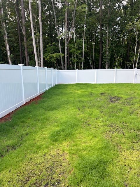 A white vinyl fence borders a green grassy backyard with a dense forest line in the background.