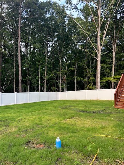 A backyard with a green lawn, a white vinyl privacy fence, and a wooded area in the background