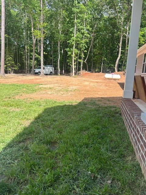 A view of a grassy yard with bare red dirt leading to a forest, with a white van and propane tank in the background.