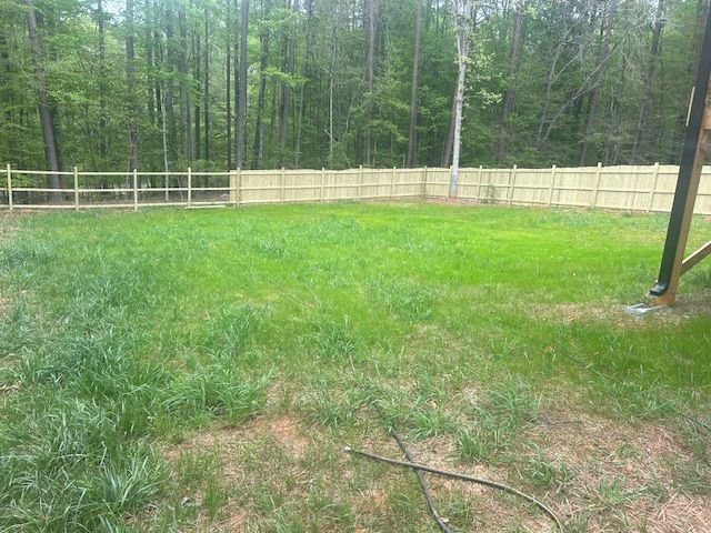 A backyard lawn with patchy green grass, bordered by a wooden fence with a line of trees in the background.