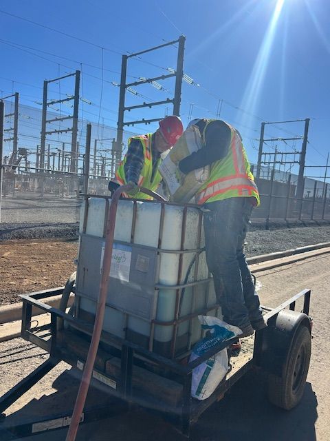 Two workers in high-visibility vests pour a bag of white powder into a large water container on a trailer at a substation.