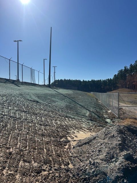 A steep, newly hydroseeded embankment slopes toward a chain-link fence under a bright blue sky.