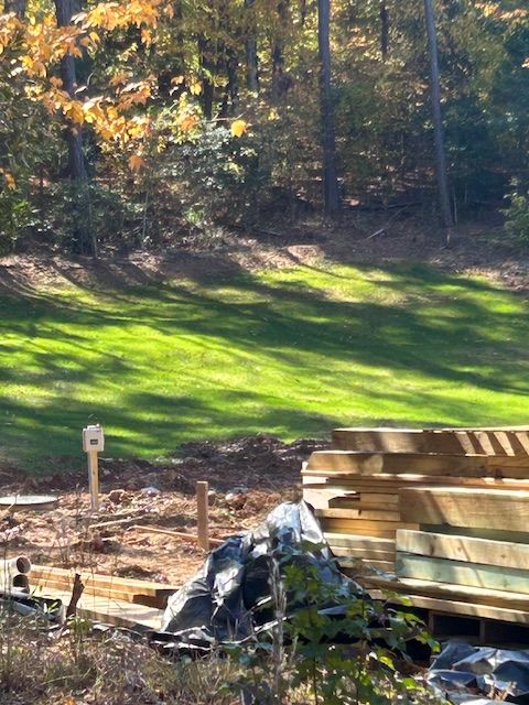 A construction site with stacked lumber piles in the foreground and a grassy, wooded hill in the background.