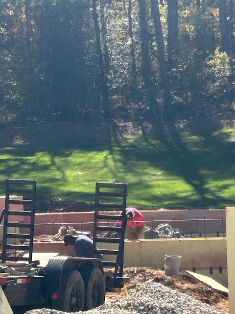 Two people work on a stone retaining wall at a construction site in front of a wooded area.