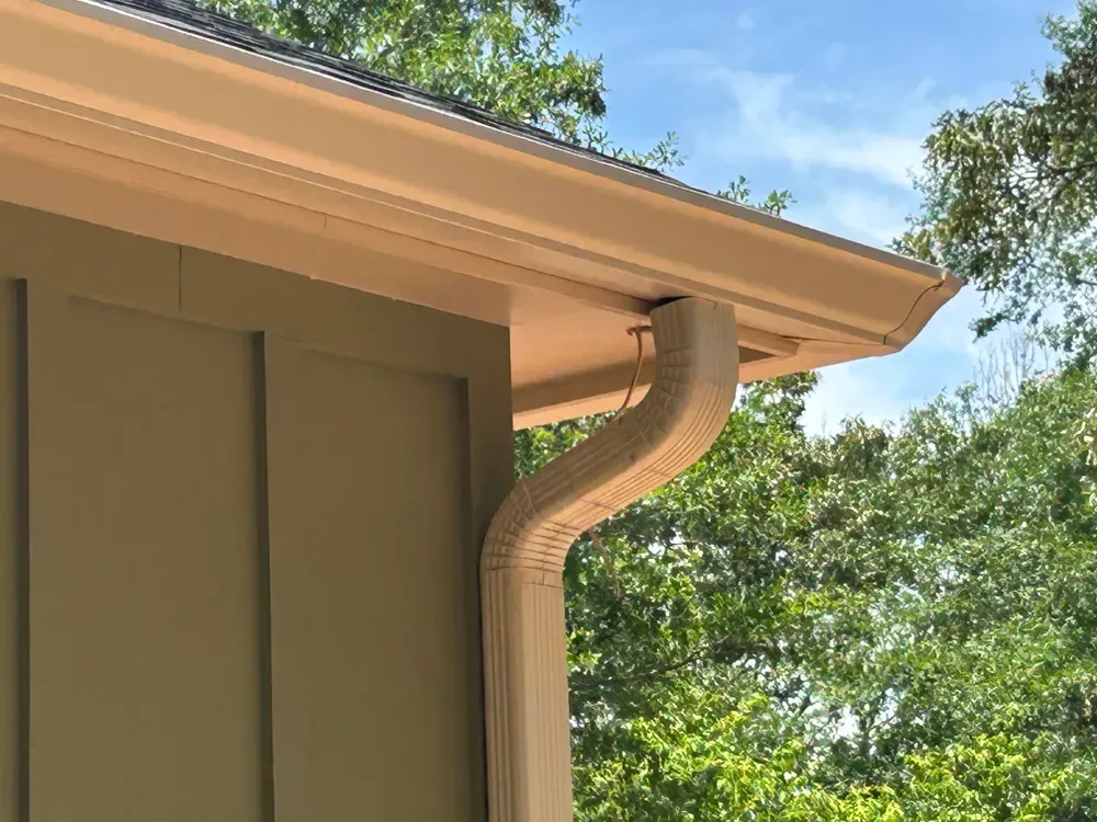 Beige gutter and downspout on a house with a green exterior, against a blue sky with trees.