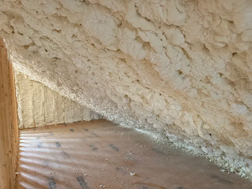 Attic interior insulated with thick, expanding foam; walls and ceiling covered in beige, textured material.
