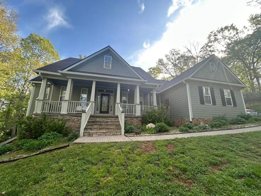 Green house with a porch, stone foundation, and a grassy yard under a blue sky.